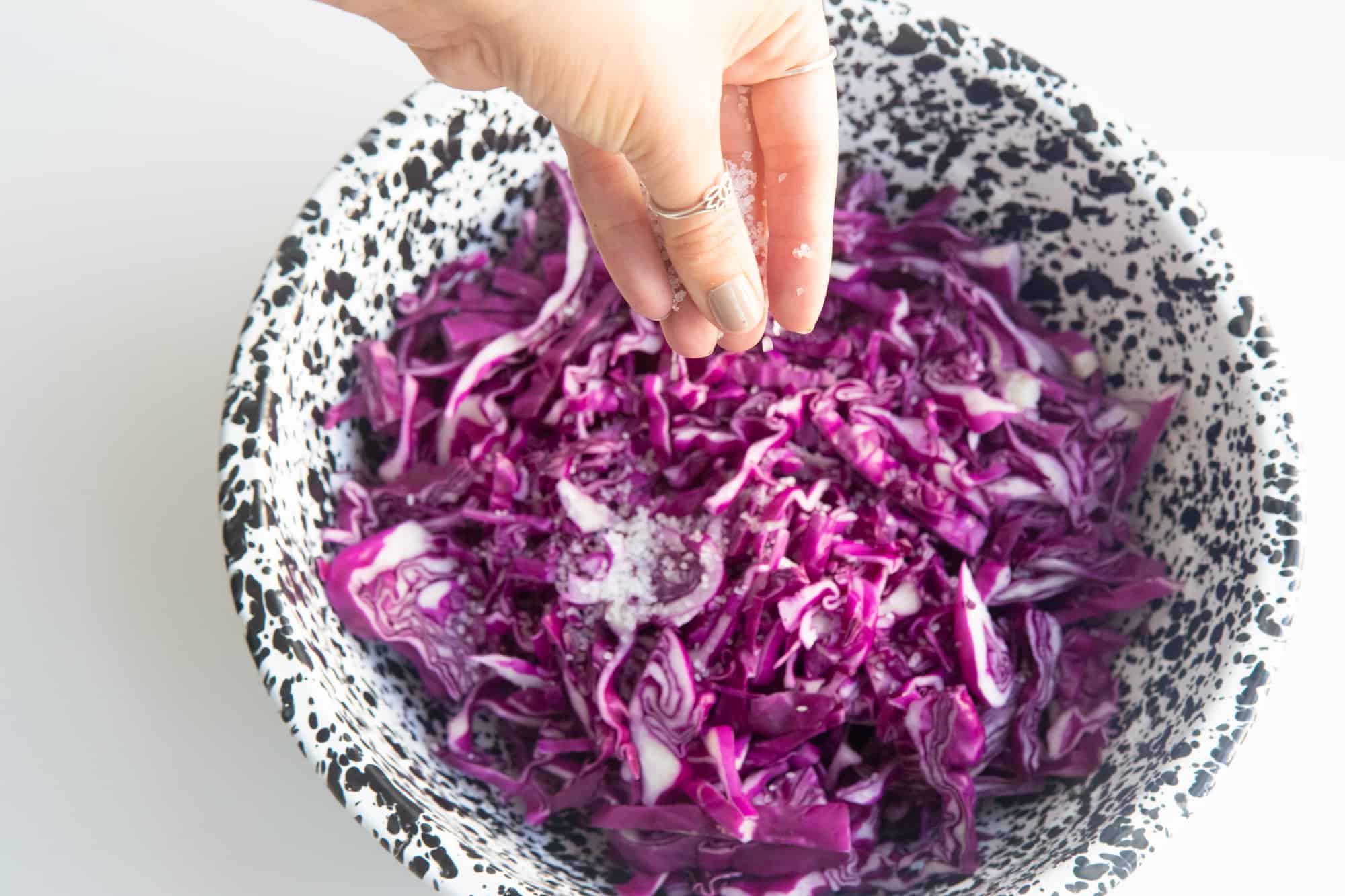 A hand sprinkles salt over a bowl of sliced red cabbage.