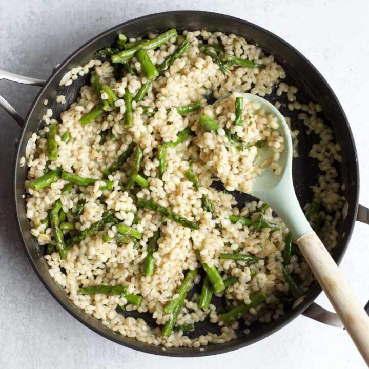 Overhead of a cooking spoon resting in a skillet of barley and asparagus.