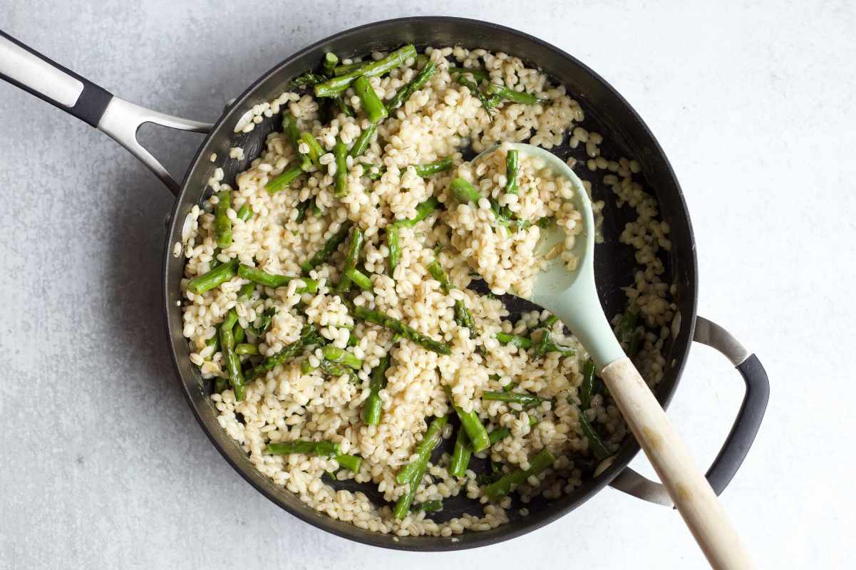 Overhead of a cooking spoon resting in a skillet of barley and asparagus.