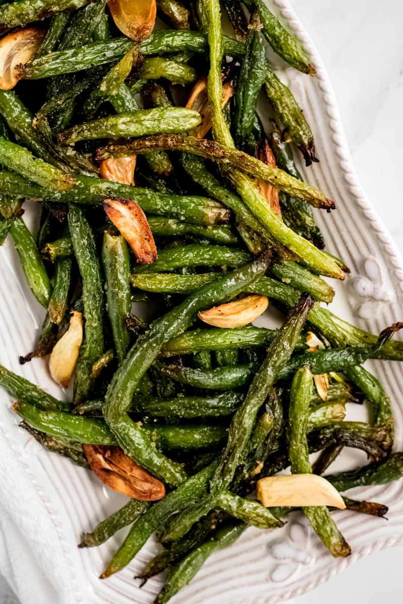 Close view of a white serving platter piled high with seasoned grilled green beans and garlic cloves.