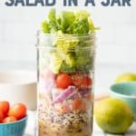 A glass jar layered with dressing, rice, beans, pico de gallo, onions, tomatoes, cilantro, and lettuce sits on a folded dish towel. Extra ingredients are scattered around the jar. A text overlay reads "Black Bean Fiesta Salad in a Jar."