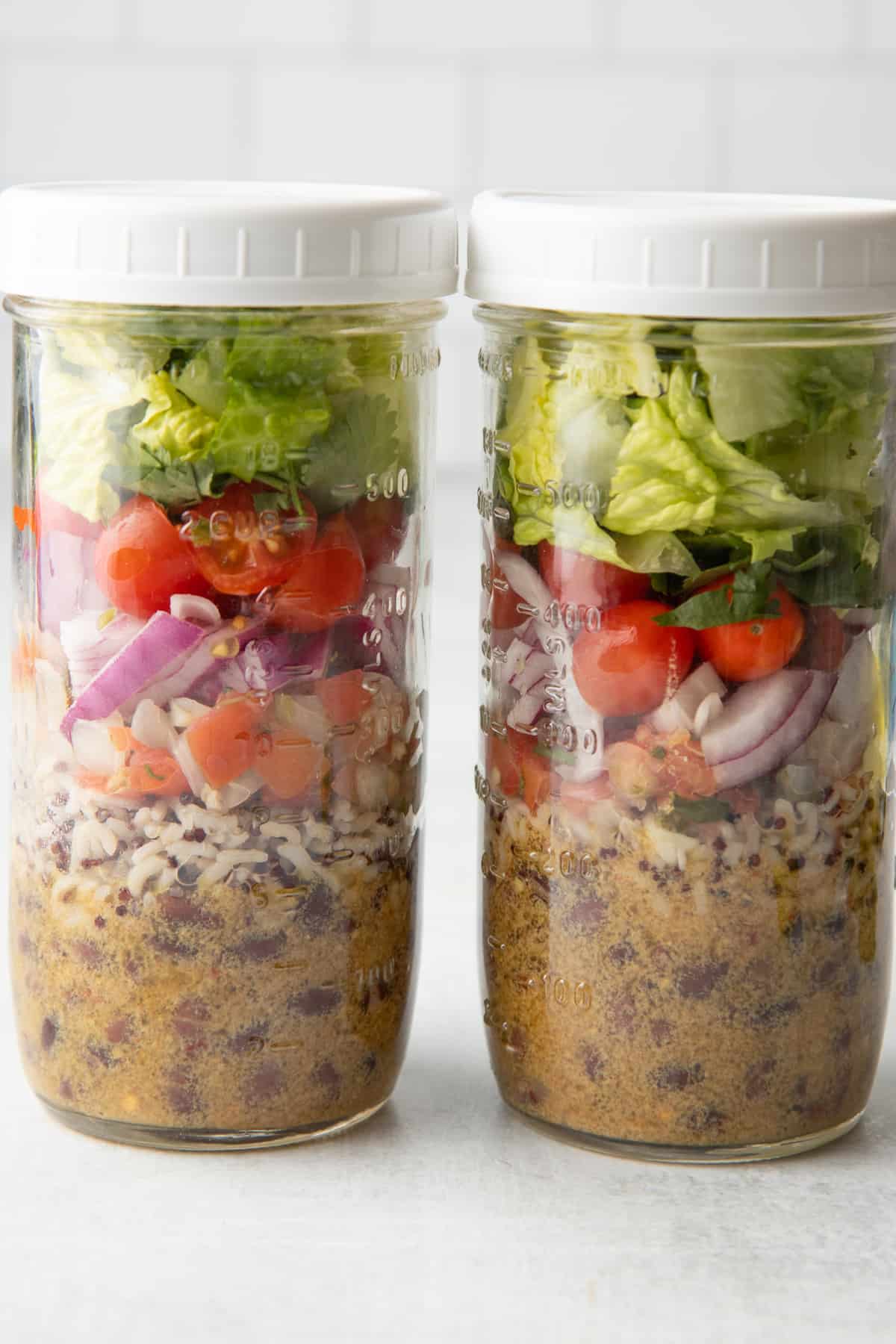 Two tall mason jars sit on a white countertop. The jars are layered with ingredients for black bean fiesta salad. Jars are topped with a white lid.