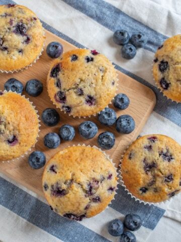 Top view of blueberry muffins on a wooden serving board with fresh berries and a blue and white striped kitchen linen.