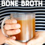 A woman's hands hold a clear mug full of bone broth with her black and white stripe shirt in the background. A text overlay reads "how to make bone broth"