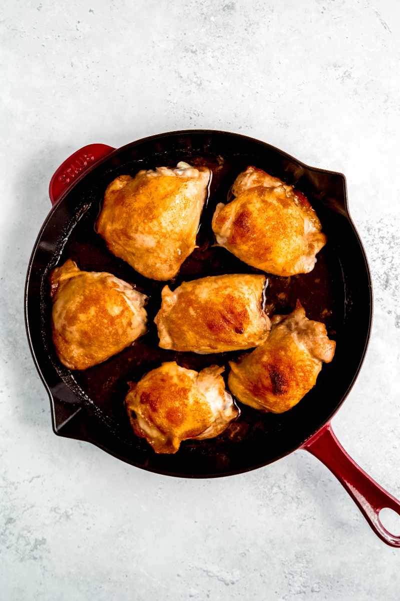 Top view of a red enameled cast iron skillet with six brown sugar chicken thighs nestled in it.