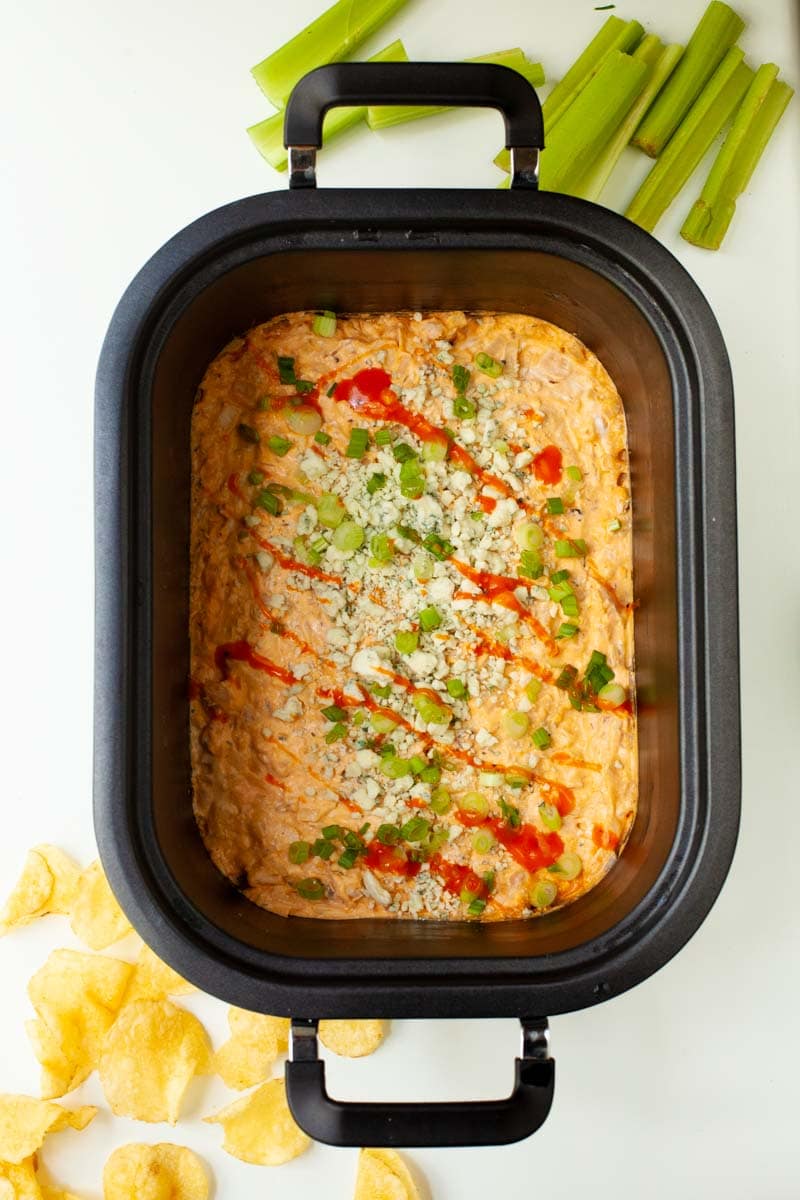 Overhead of a slow cooker filled with homemade dip garnished with blue cheese, chopped scallions, and a buffalo sauce drizzle.