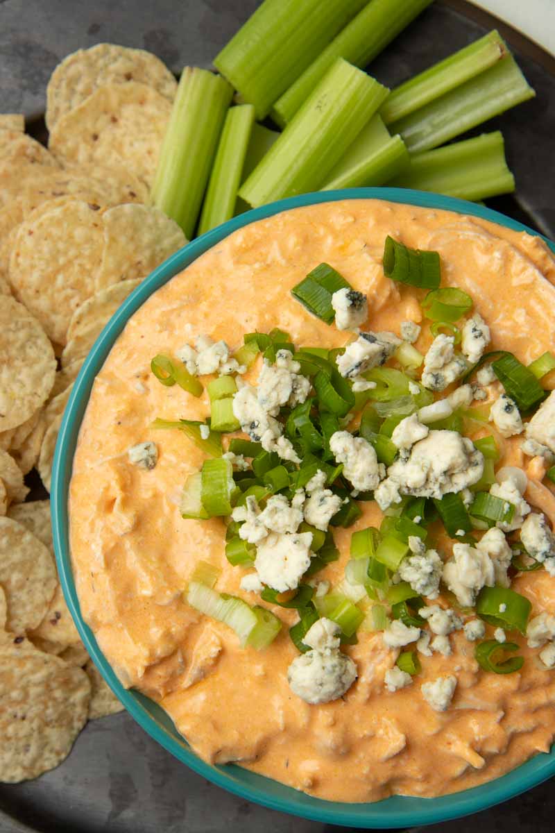 A serving bowl of Instant Pot buffalo chicken dip rests on a metal platter with tortilla chips and celery sticks beside it.