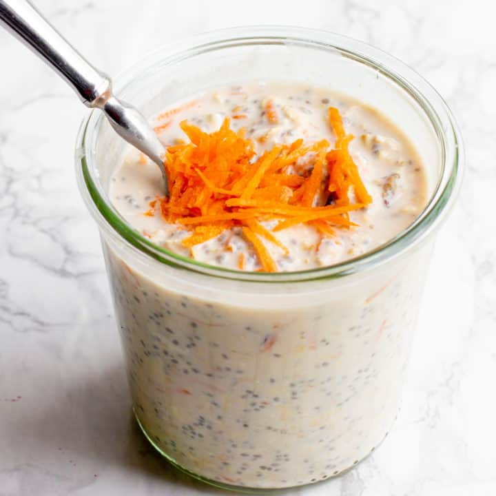 A glass jar filled with carrot cake overnight oats sits on a marble countertop. The oats are garnished with shredded carrots, and a spoon dips into the jar.