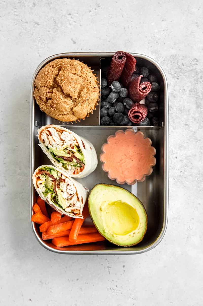 Overhead of a metal bento-style lunchbox packed with chicken bacon ranch wraps, fruit, veggies, and a muffin.