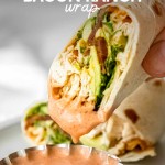 A hand dips a chicken bacon ranch wrap cut on the bias into a small scalloped bowl of spicy ranch dip. A text overlay reads, "Chicken Bacon Ranch Wrap."