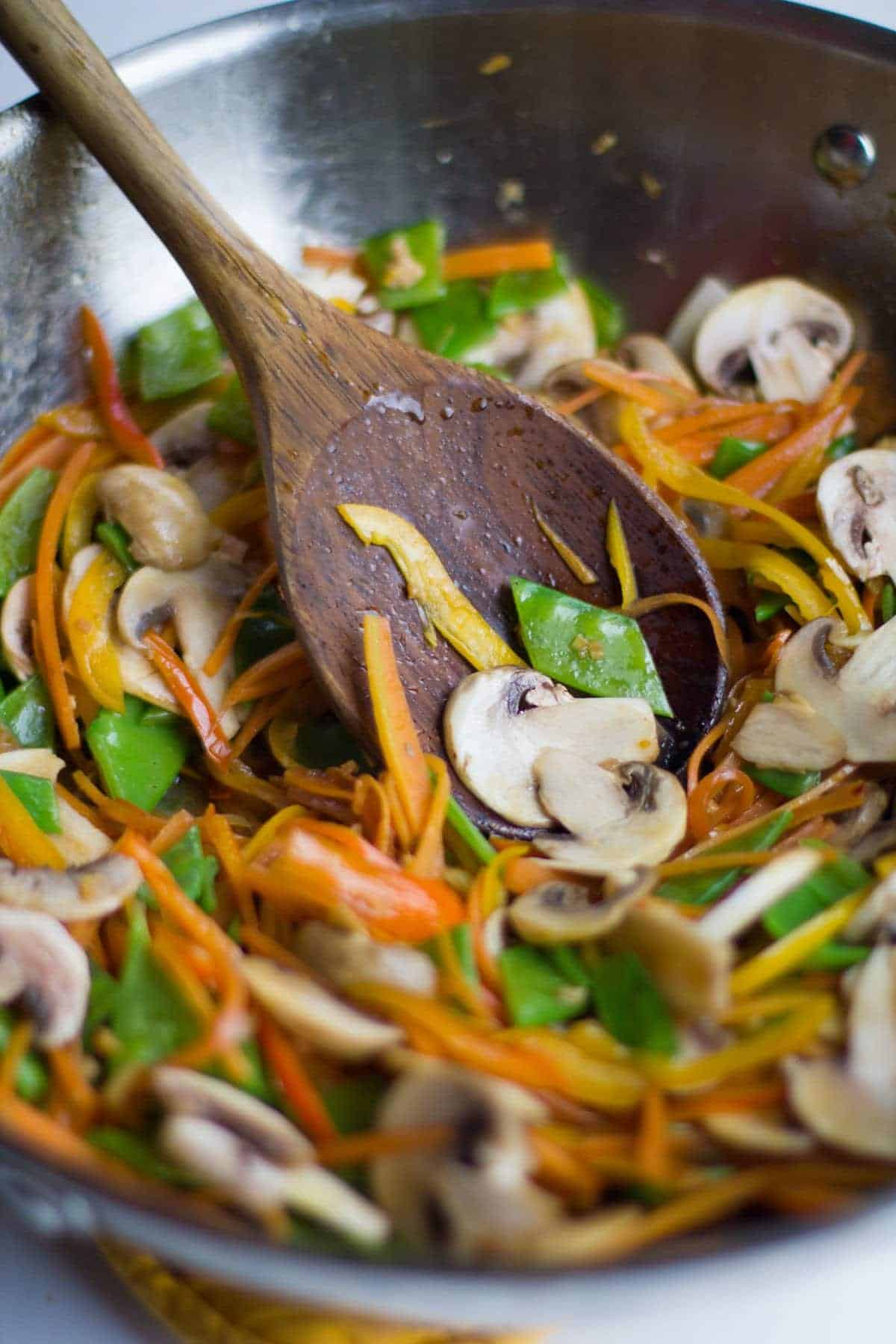 Partially-cooked vegetables get mixed with a wooden spoon in a wok.