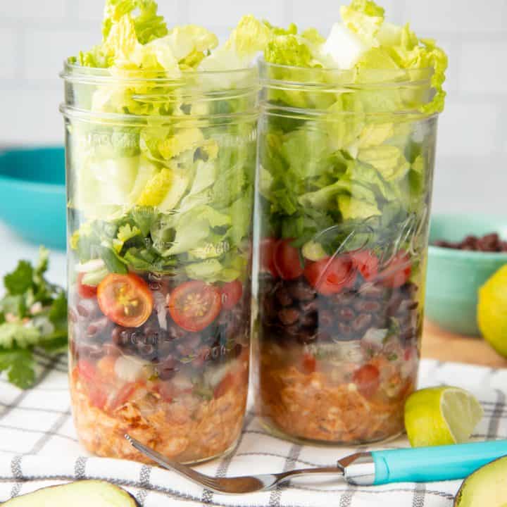 Two mason jars are layered with chicken, black beans, tomatoes, and lettuce. A fork with a teal handle rests in front of the jars.