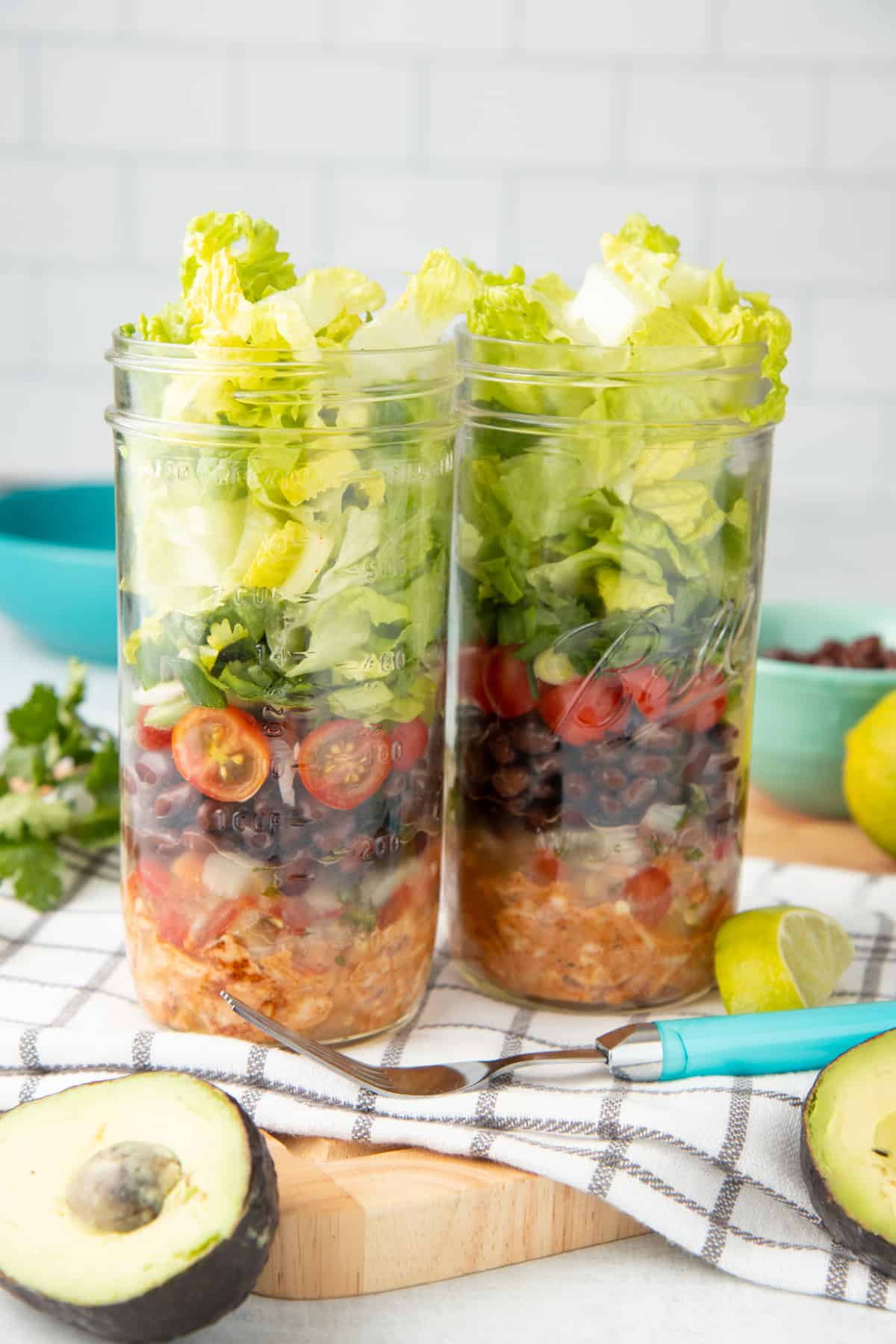 Two mason jars are layered with chicken, black beans, tomatoes, and lettuce. A fork with a teal handle rests in front of the jars.