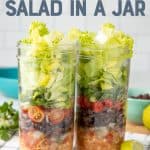 Two mason jars are layered with chicken, black beans, tomatoes, and lettuce. A fork with a teal handle rests in front of the jars. A text overlay reads "Chicken Taco Salad in a Jar."