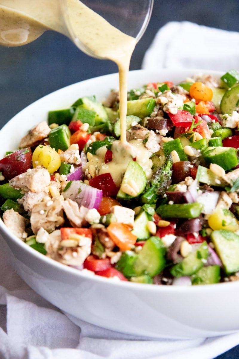 Side angle shot of Grilled Chicken and Asparagus Chopped Greek Salad in a white bowl, with dressing being drizzled over the salad