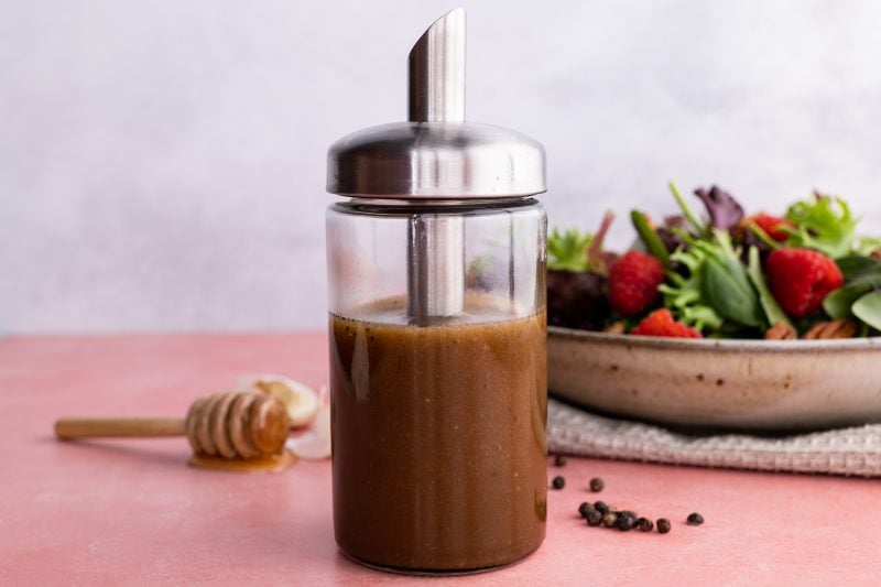 Close view of homemade balsamic vinaigrette in a glass cruet in front of a fresh salad.