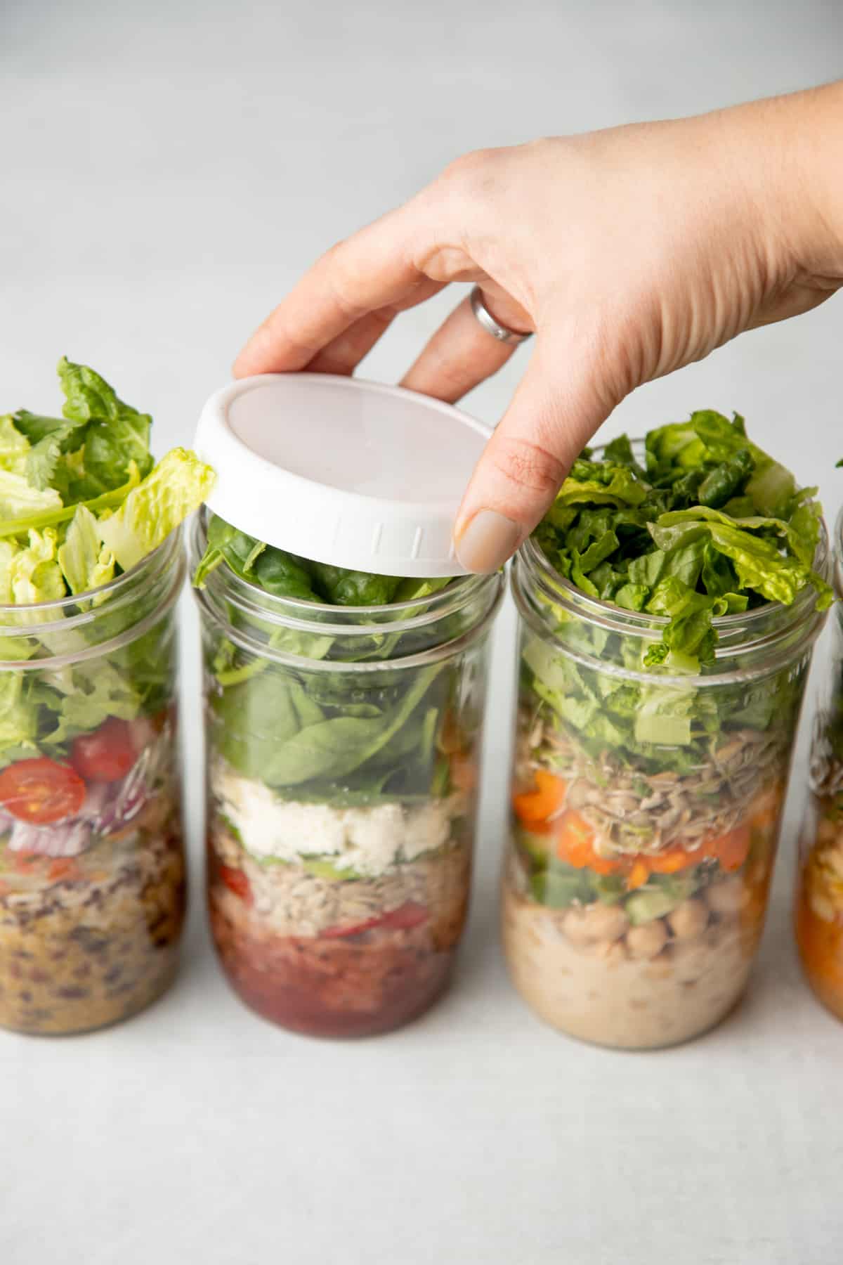 Three tall mason jars are layered with salad ingredients. A hand places a white lid on the middle jar.