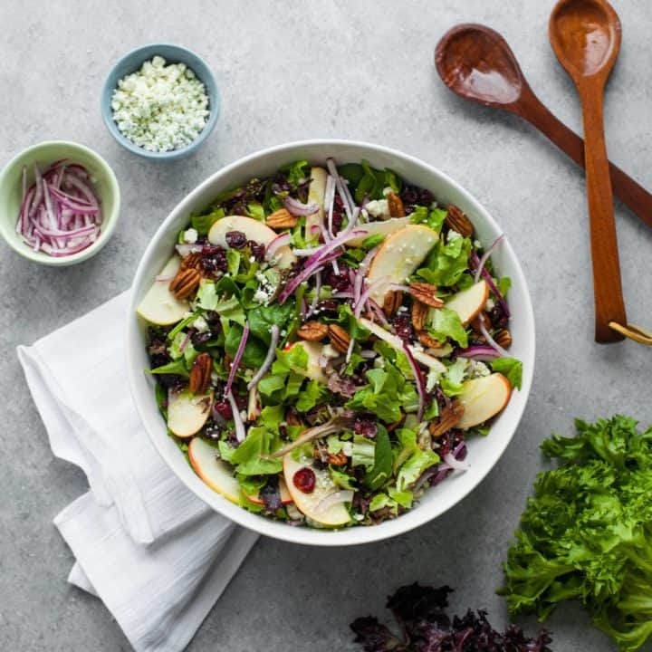 Overhead of cranberry apple pecan salad in a white serving bowl, with wooden serving spoons and fresh ingredients around it.