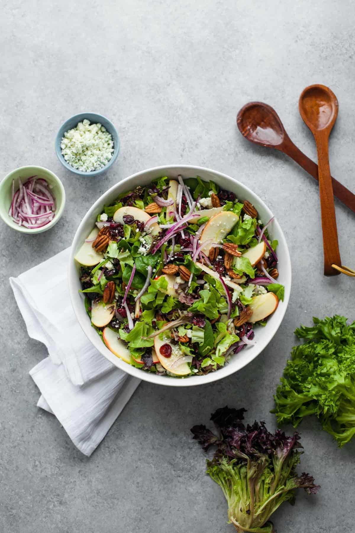 Overhead of cranberry apple pecan salad in a white serving bowl, with wooden serving spoons and fresh ingredients around it.