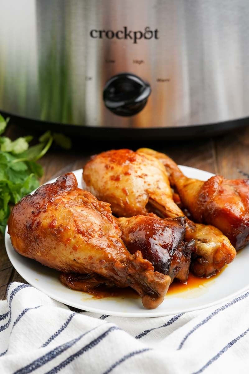 A platter of slow cooked chicken drumsticks on a white plate in front of a Crockpot.