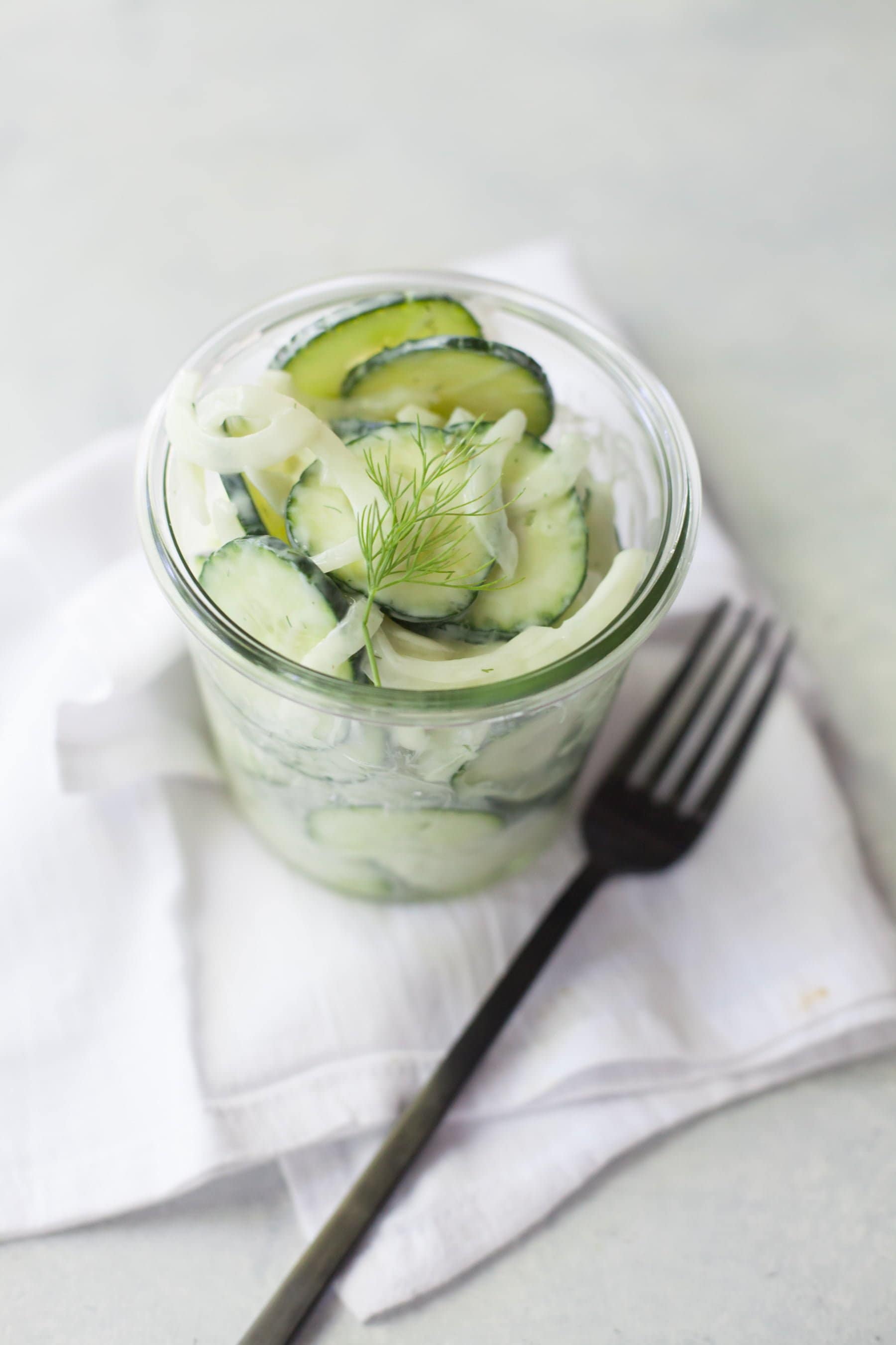 Cucumber salad in a jar on a white cloth napkin with a fork beside it.