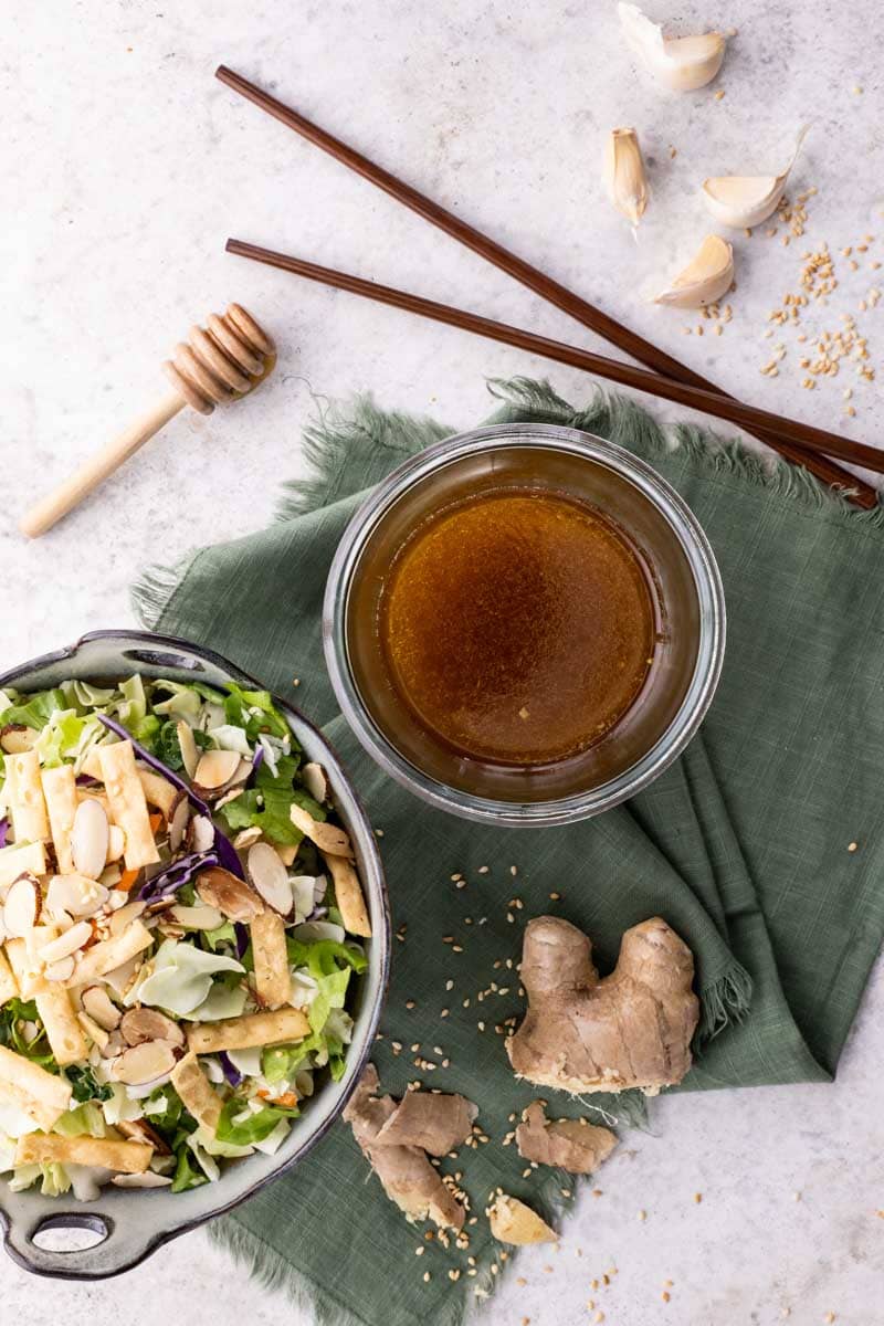 Overhead of sesame ginger dressing in a bowl beside a bowl of salad, fresh ginger, garlic, and sesame seeds around them.