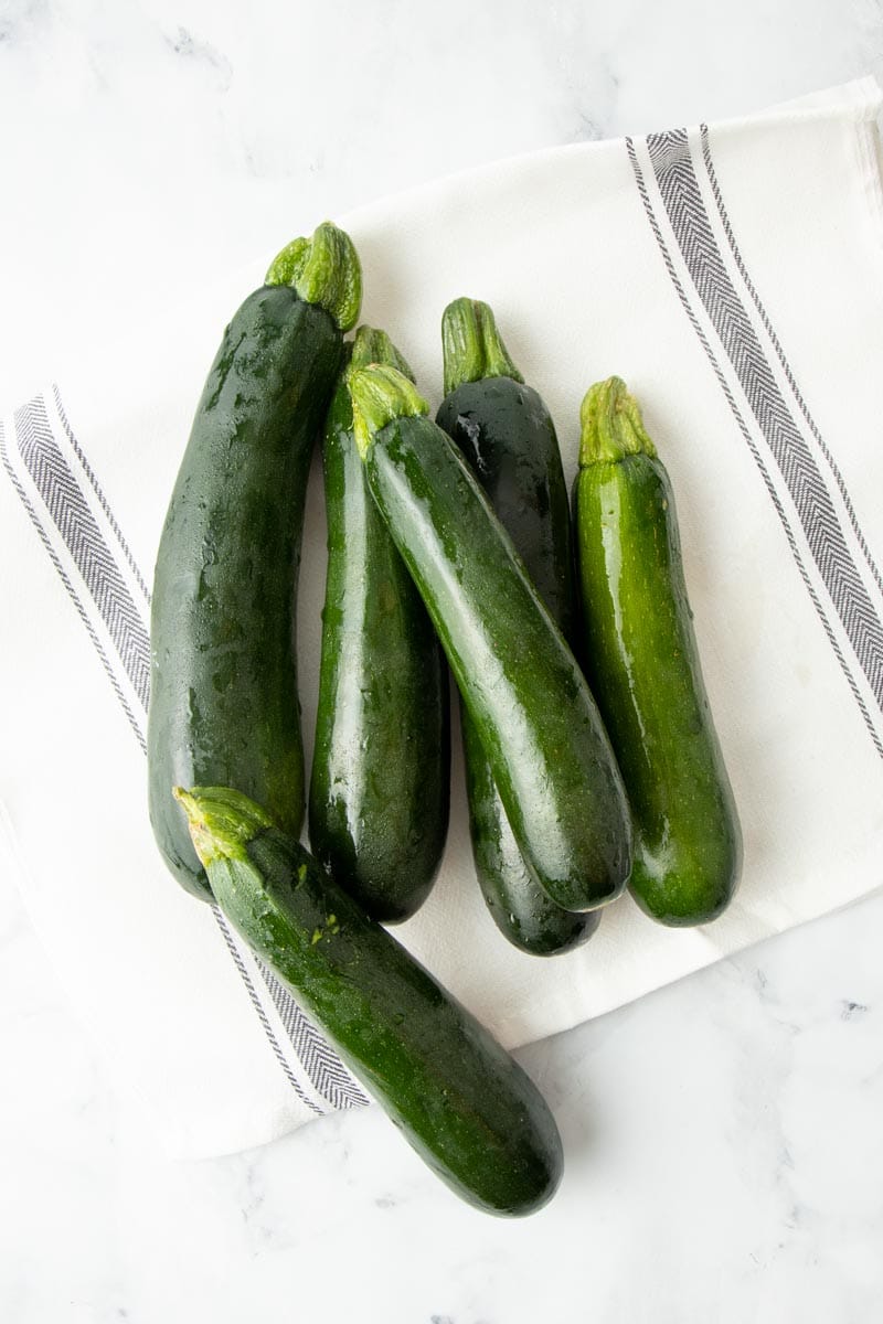 A pile of fresh zucchini still wet from washing sits atop a kitchen towel on a marble countertop.