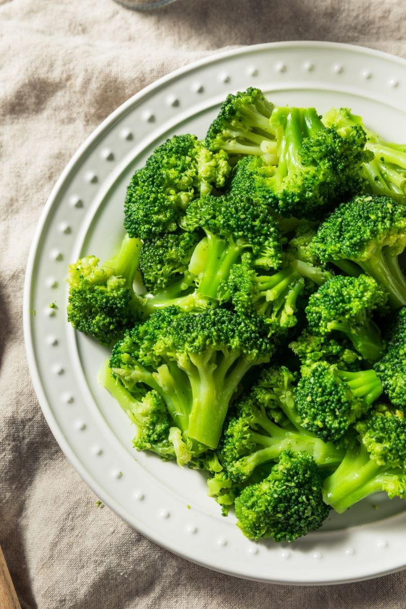 Overhead view of a pile of garlic broccoli on a white plate, a linen tablecloth beneath it.