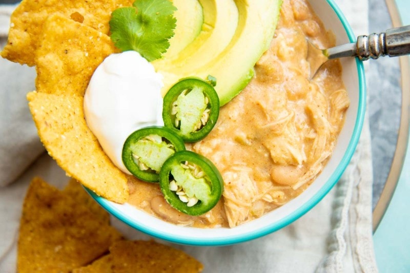 Close view of creamy white chili topped with jalapeno slices, sour cream, avocado, and corn tortilla chips.