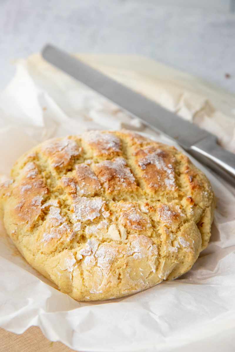 A crusty loaf of gluten-free yeast bread rests on parchment paper with a bread knife beside it.