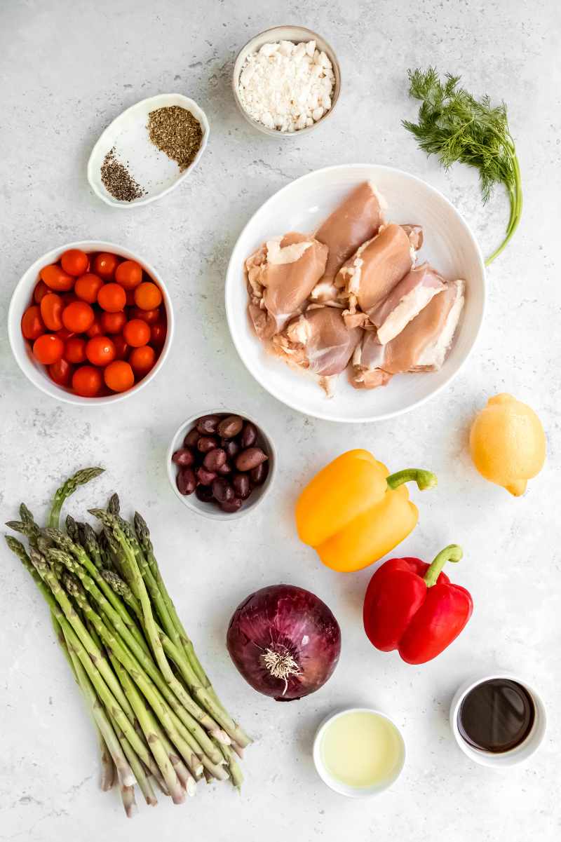 Overhead of ingredients including chicken thighs, kalamata olives, bell peppers, asparagus, red onion, and lemon.