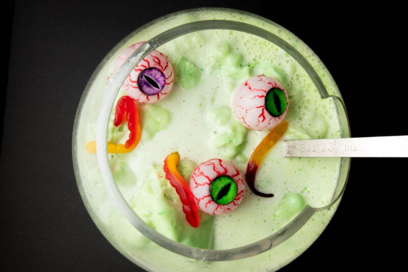 Overhead of a ladle in a large punch bowl filled with Halloween punch garnished with gummy worms and plastic eyes on a black background.