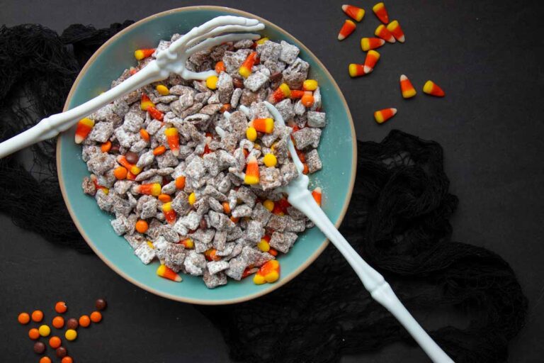 Overhead of Halloween puppy chow served in a teal bowl on a black cloth, two skeleton hands reaching into the bowl.