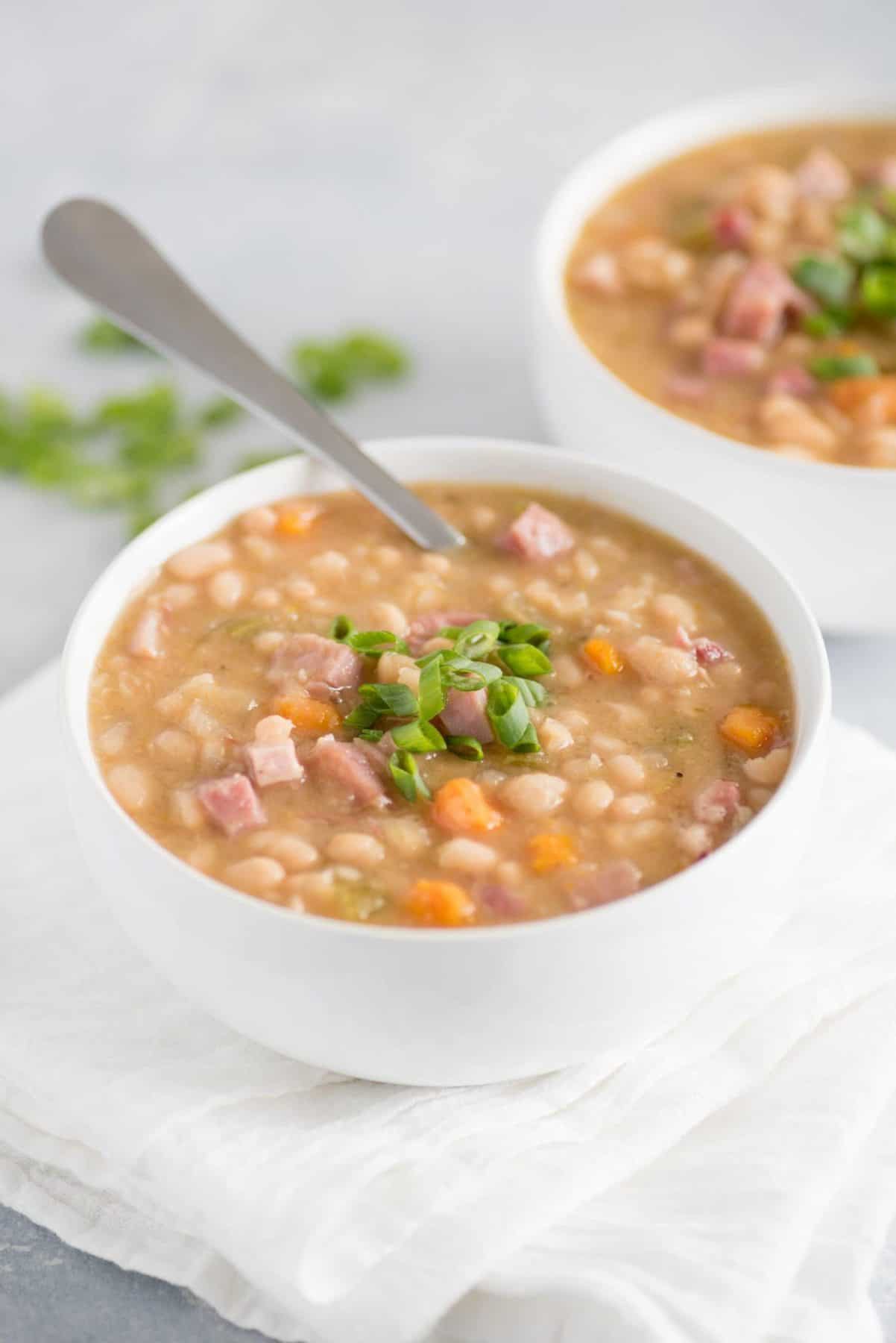 Cooked ham and beans in a white bowl with a spoon and scallion garnish.