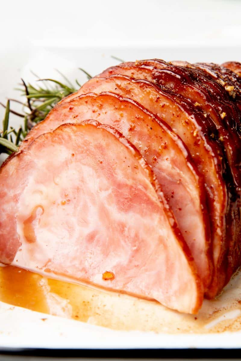 Ham sits on a white dish. Juices from the glaze surrounds the ham.