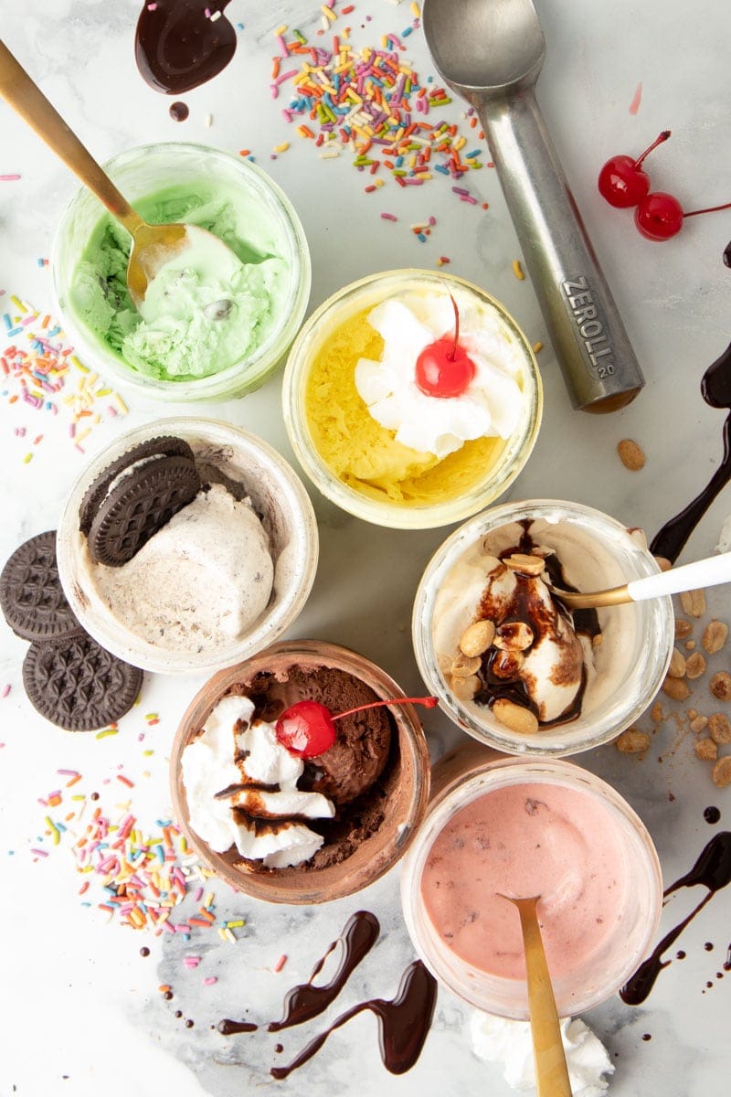 Overhead shot of various flavors of mason jar ice cream, with various toppings around the jars. Flavors include strawberry, peanut butter, cookies n cream, lemon, chocolate, and mint chocolate chip.