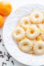 Clementine and Clove Spritz Cookies on a white plate with clementines and cloves nearby