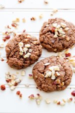 Three chewy nutella cookies topped with chopped hazelnuts on a white wooden table.