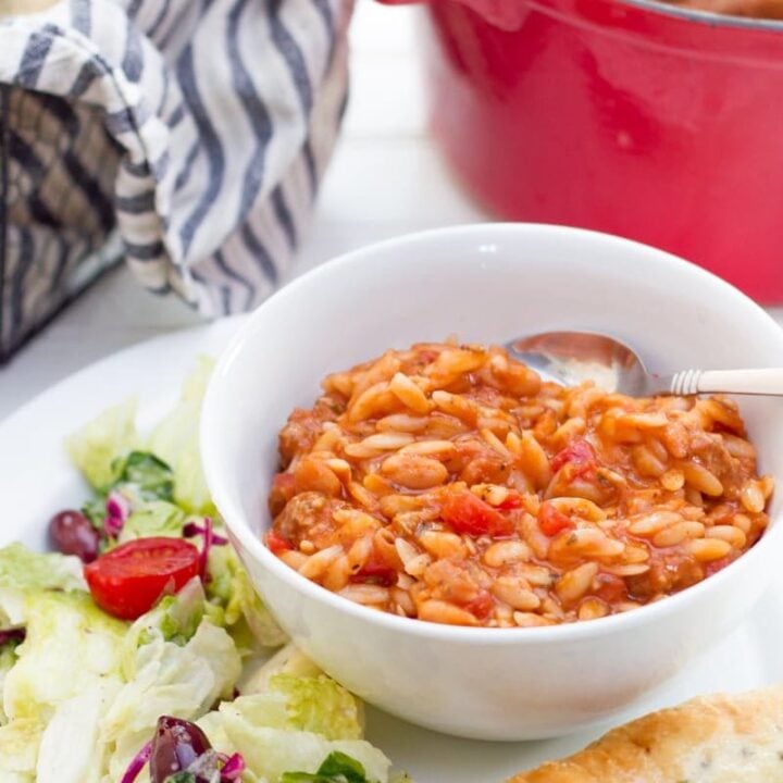 A bowl of Pasta e Faglioli sits on a white plate, surrounded by lettuce and a breadstick. A red dutch oven is seen in the background.