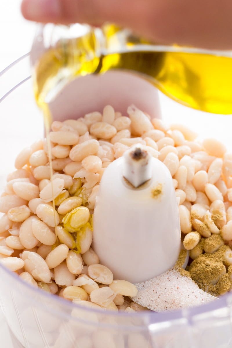 Olive oil streams into a food processor that already holds white beans.