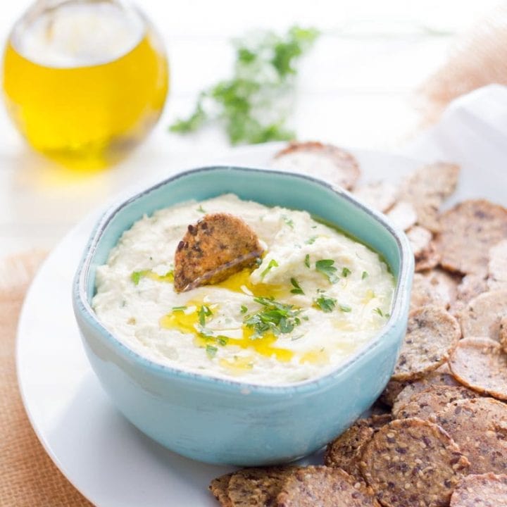 White bean dip drizzled with olive oil is in a light blue bowl, surrounded by crackers on a white platter.