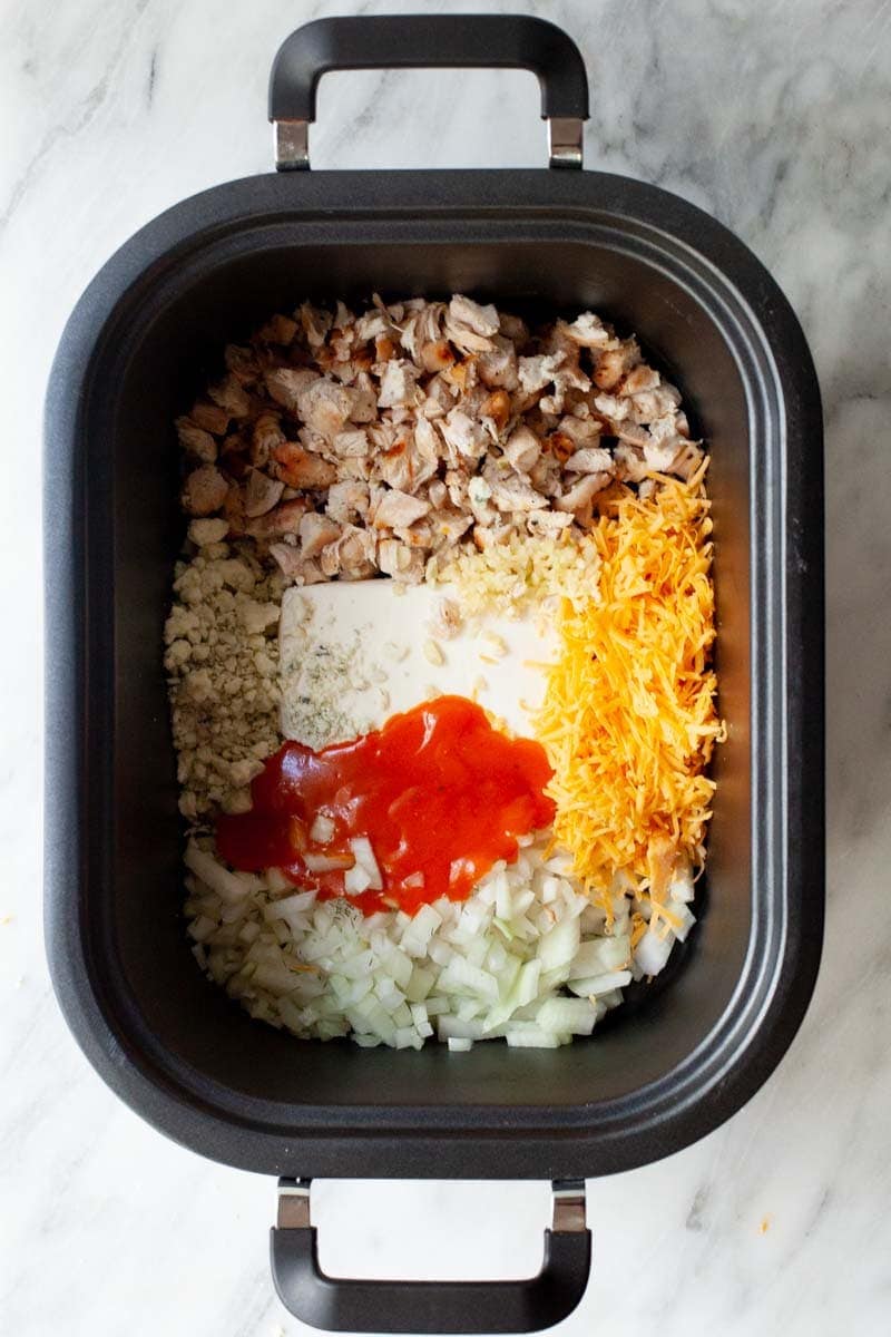 Ingredients such as chicken, cheese, and buffalo sauce in the basin of a slow cooker