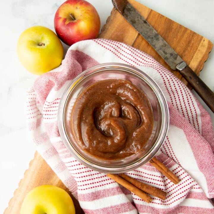 A jar of finished apple butter stands with cinnamon sticks and whole apples around it.