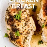 Close view of a juicy chicken breast on a plate in flavorful juices with fresh herb garnish. A text overlay reads, "Instant Pot Chicken Breast."