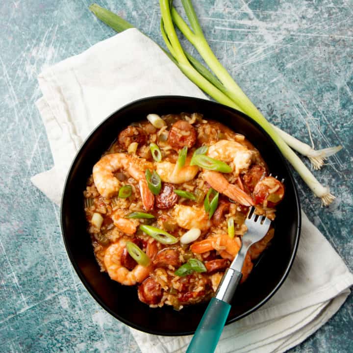 A piece of sausage rests on a fork inside a bowl of jambalaya. Whole green onions lay alongside the bowl.