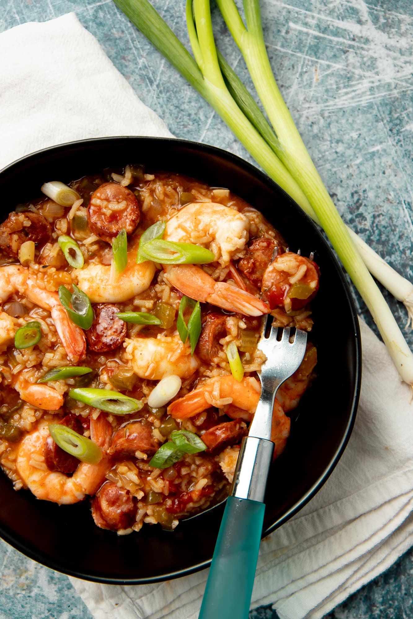 A piece of sausage rests on a fork inside a black bowl of shrimp jambalaya. The bowl is on a white napkin on a teal background.