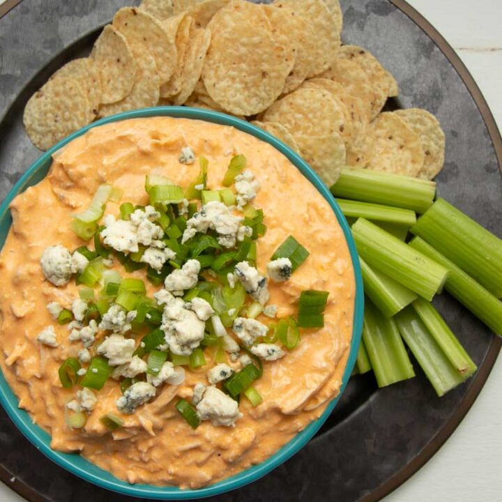 Overhead of a serving platter with a teal bowl of Instant Pot buffalo chicken dip surrounded by round tortilla chips and celery sticks.