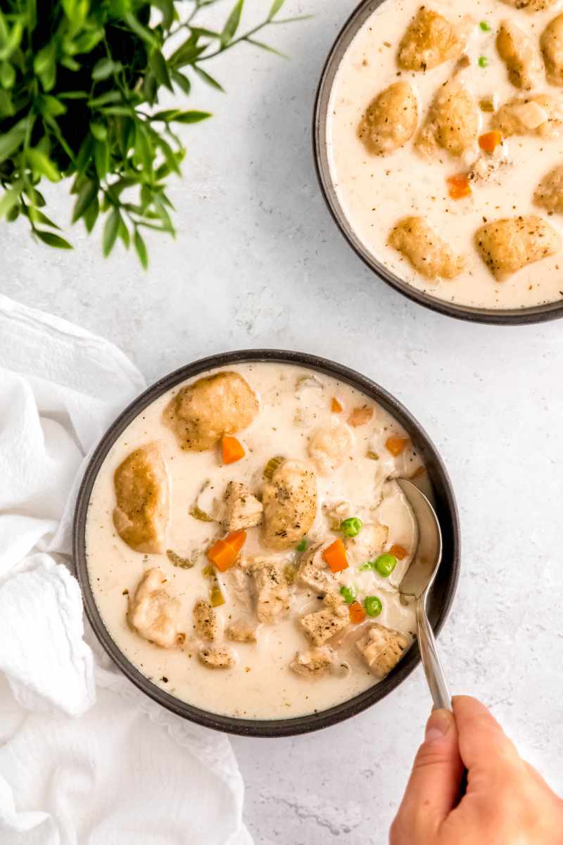 A hand dips a spoon into a bowl of chicken soup with dumplings.