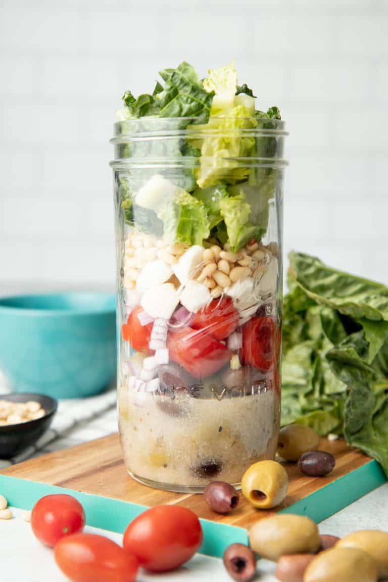 A mason jar salad sits on a teal-edged cutting board, surrounded by lettuce, olives, and cherry tomatoes.