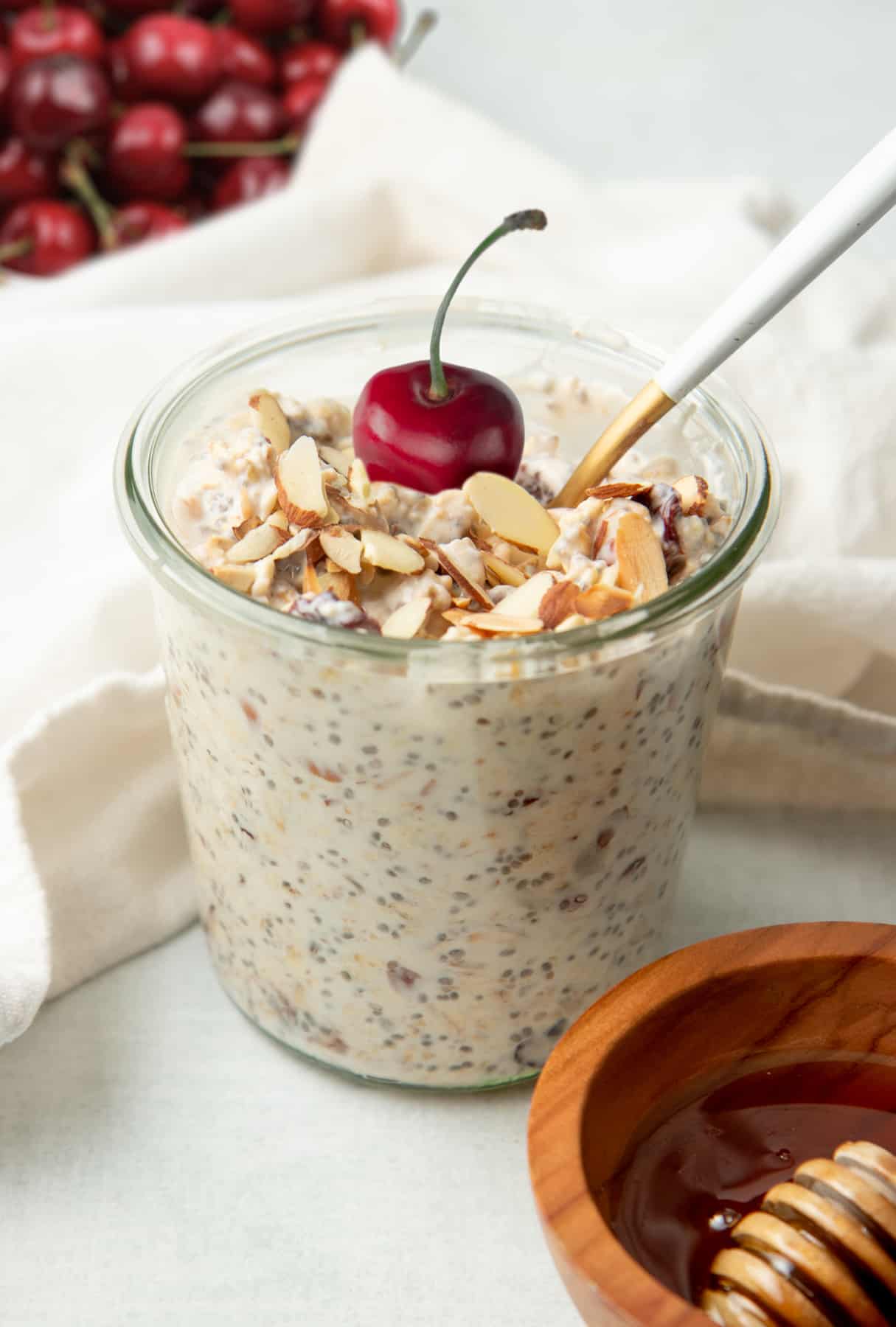 A spoon rests in a glass jar of cherry and almond overnight oats. The oats are garnished with a whole fresh cherry.